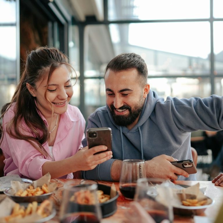 Rezension von Kunden Zwei Personen sitzen an einem Tisch, lächeln und schauen auf ein Smartphone. Essen und Getränke sind sichtbar.